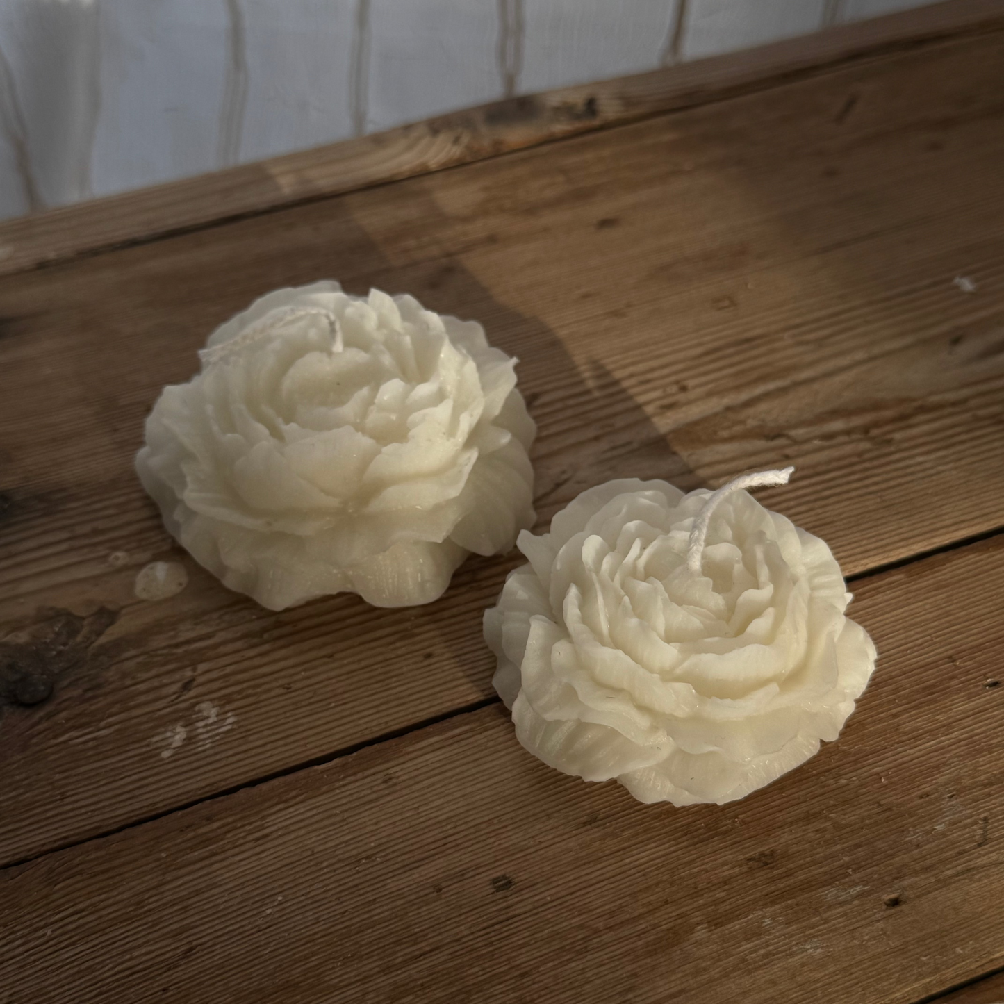 Two white rose-shaped candles on a wooden surface with a white wall background