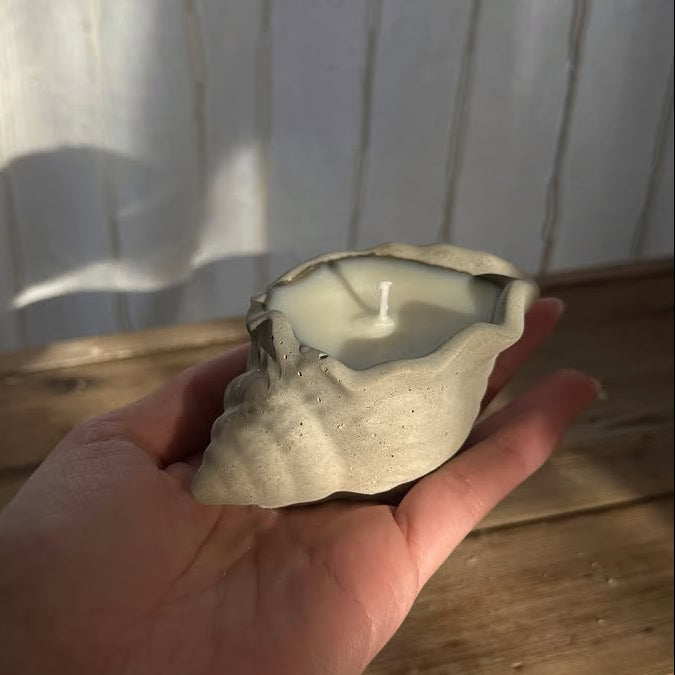 Hand holding a shell-shaped candle against a wooden surface and white curtain background